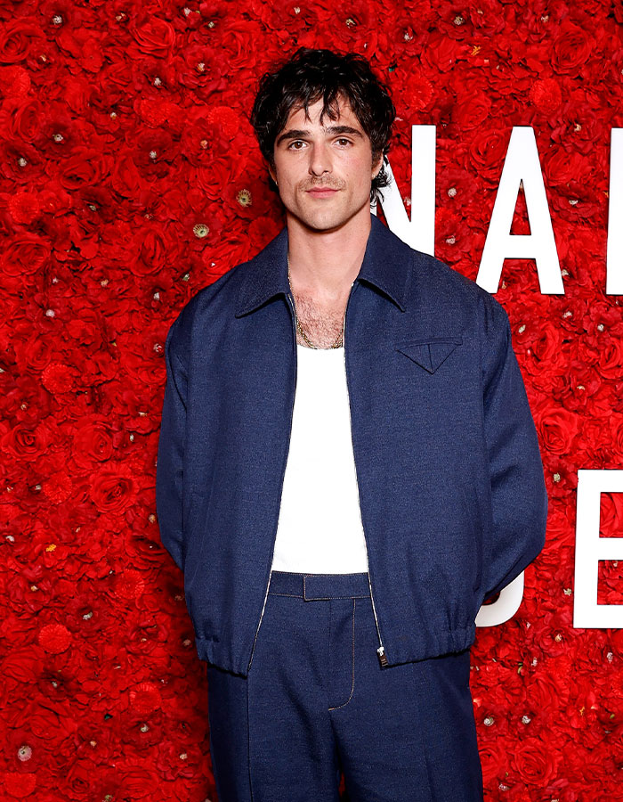 Actor posing in navy outfit against a red rose wall backdrop at event for controversial Wuthering Heights adaptation.