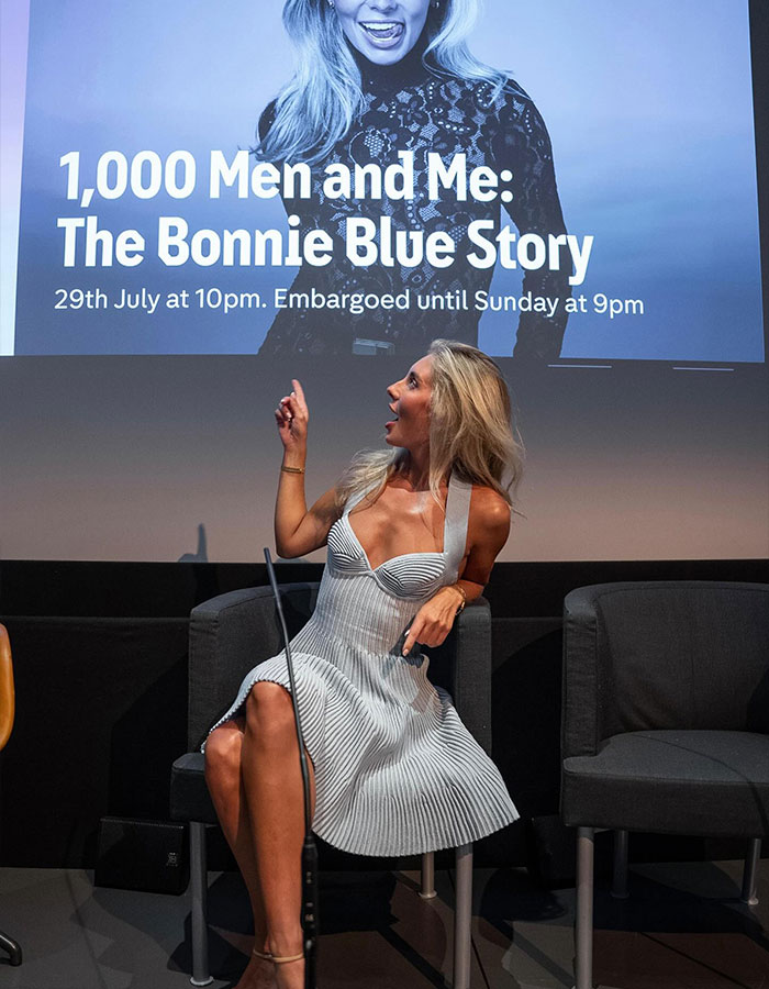 Woman in a white dress seated on stage, pointing up at a screen about Bonnie Blue&rsquo;s career in a new documentary.