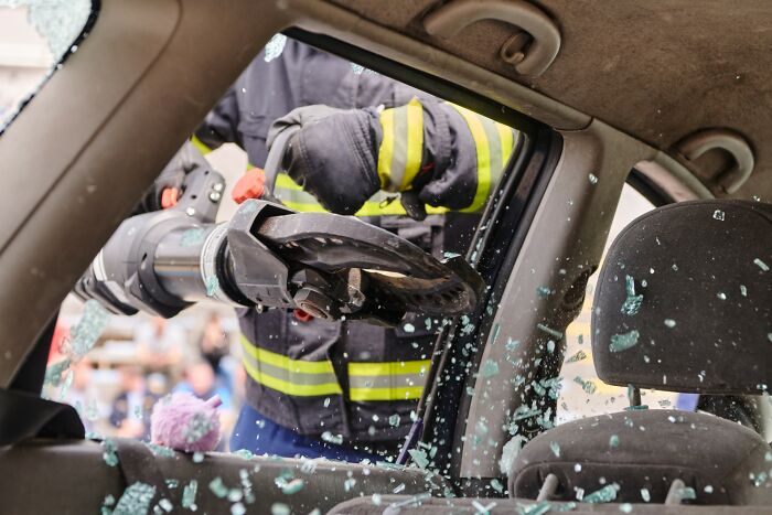 Firefighter using hydraulic rescue tool to break a car window in an emergency accident rescue scene, related to autopsy tech risks. - 13