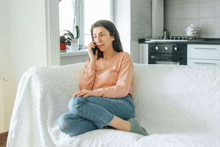 Woman sitting on white couch at home, talking on phone, illustrating woman correcting mother-in-law about baby&rsquo;s name.