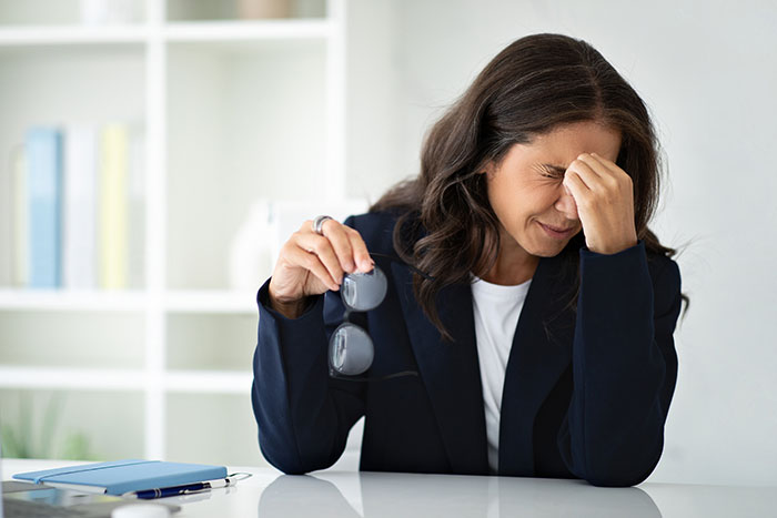 Woman stressed at desk holding glasses with eyes closed, reacting to unexpected email and video file on computer.