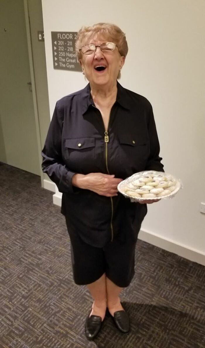 Elderly woman smiling warmly, holding a plate of cookies, embodying wholesome kind neighbors in a residential hallway.