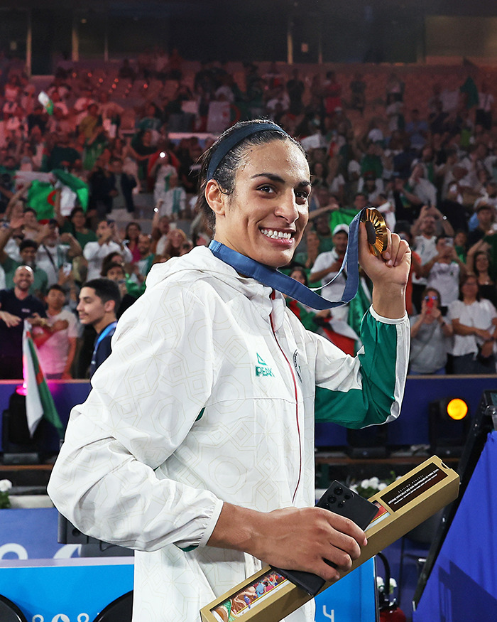 Imane Khelif holding a gold medal and award, smiling at a crowded sports event celebrating a dramatic makeover.