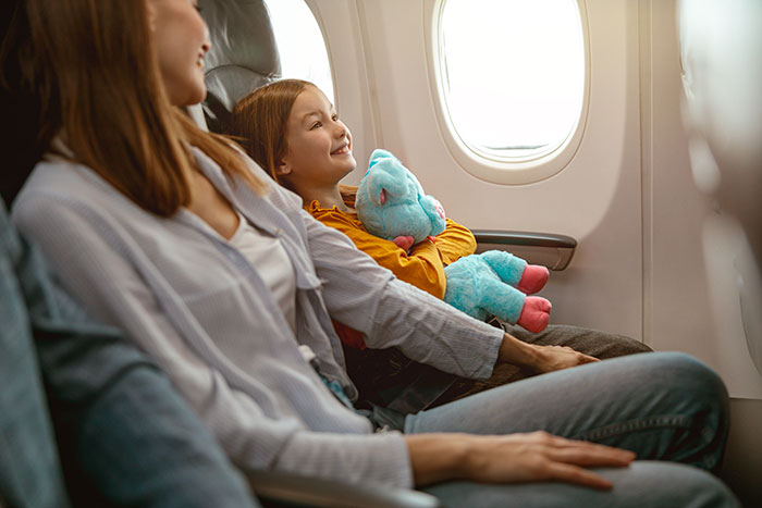 Mom and child sitting on an airplane, child holding a stuffed animal, representing a 6-leg flight experience with kids.