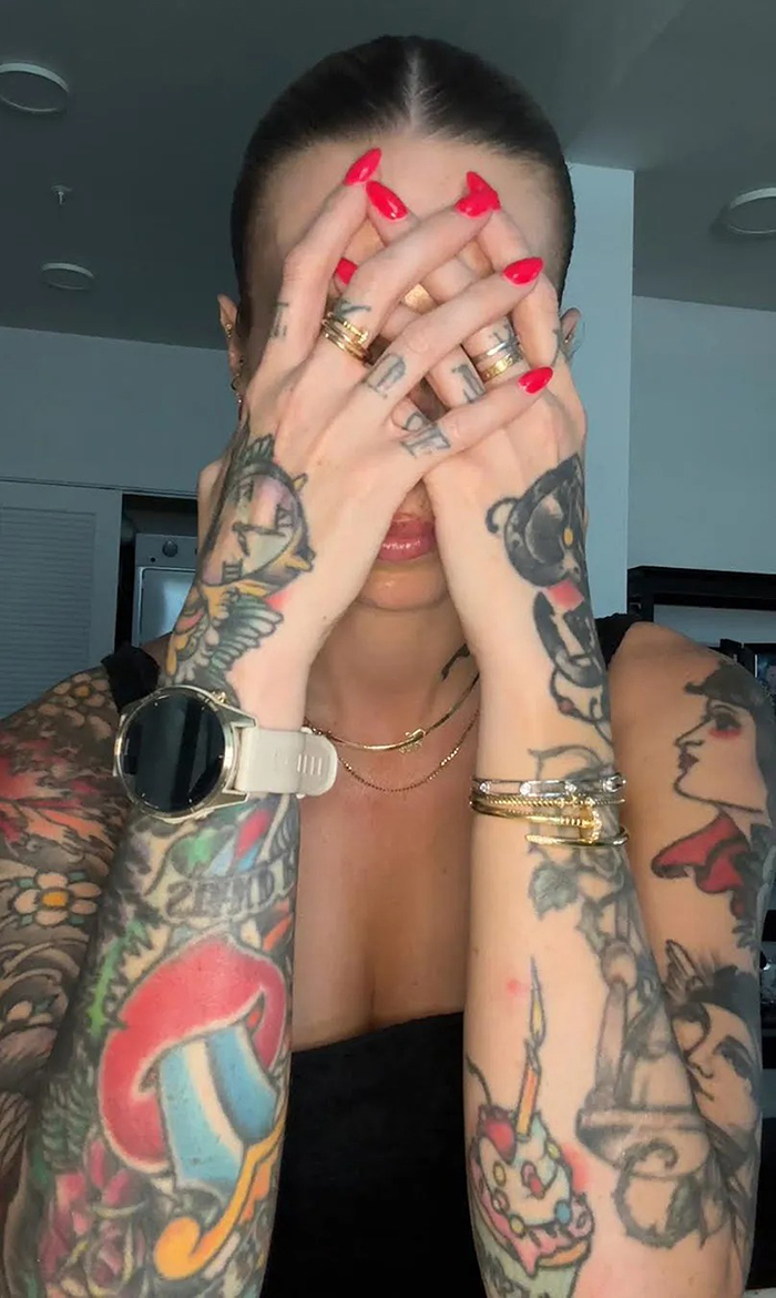 Mom with over 30 tattoos covering arms and hands, wearing gold rings and a smartwatch, hiding her face with red-painted nails.