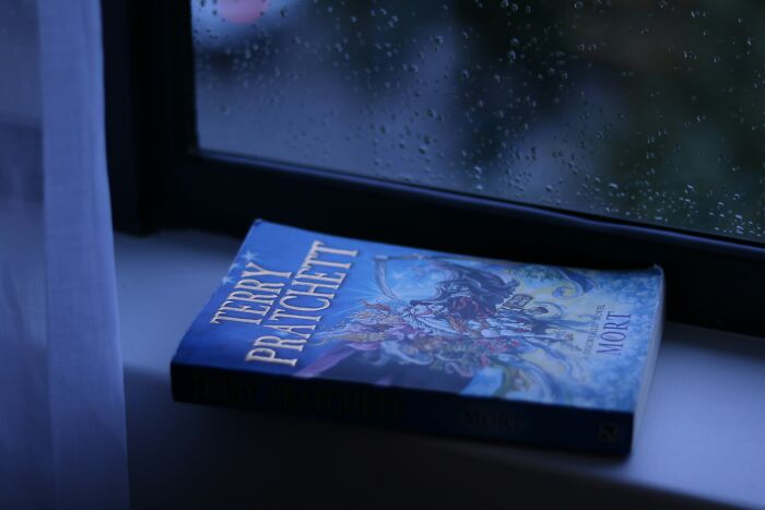 Book by Terry Pratchett on a windowsill with raindrops outside, illustrating unusual money-making hacks concept.