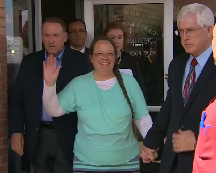 Kim Davis smiling and waving while leaving a courthouse, surrounded by several people including a man in a suit holding her hand.