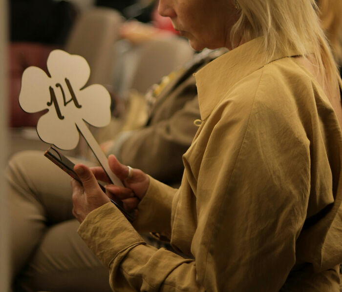 Woman in tan jacket holding a clover-shaped paddle with number 14, highlighting wealth gap perspective at an auction event.