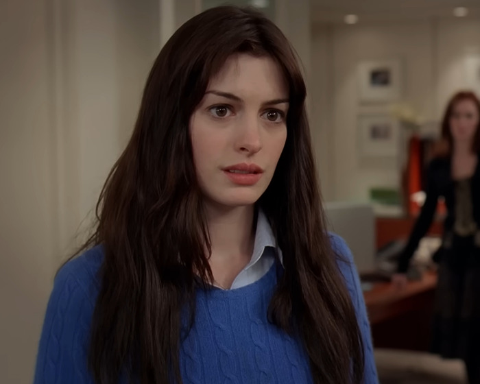 Anne Hathaway with long dark hair and blue sweater reacting with concern on a movie set interior.