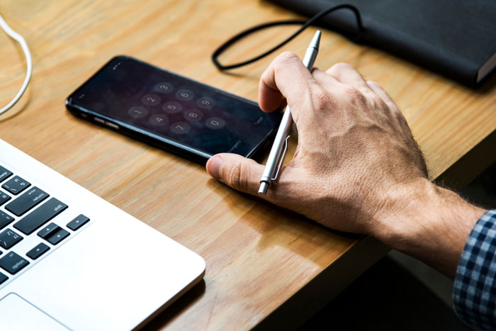 Hand holding pen over smartphone with passcode screen beside laptop on wooden desk, highlighting creepy guy incessantly texting. Hand holding pen over smartphone with passcode screen beside laptop on wooden desk, highlighting creepy guy incessantly texting.
