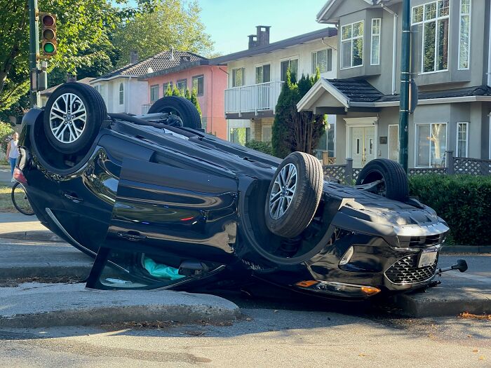 Black car flipped upside down on urban street with traffic light and residential houses, highlighting people surviving crazy situations.