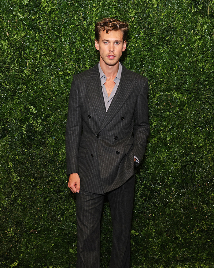 Austin Butler posing in a dark pinstripe suit against green foliage, showcasing peak male body confidence and style.