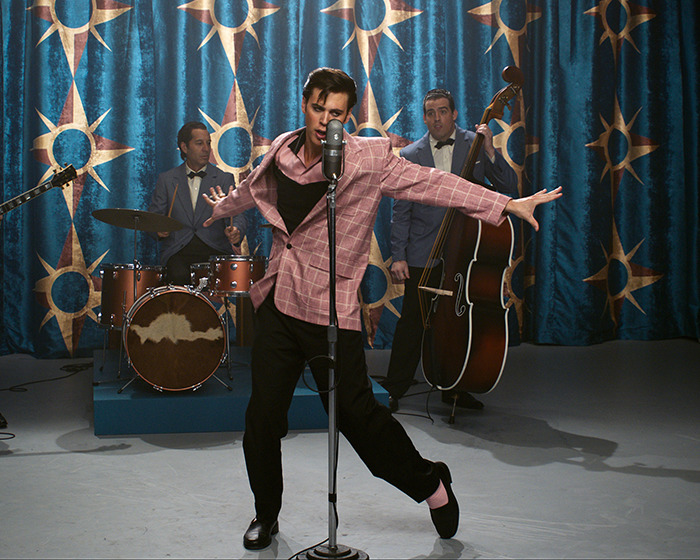 Austin Butler performing energetically in front of a vintage microphone during a band photoshoot with a retro backdrop.
