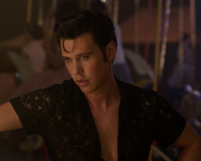 Austin Butler in a dimly lit setting wearing a black lace shirt, showcasing a peak male body look.