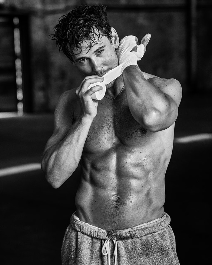 Shirtless man with peak male body wrapping hands, showing muscular physique and intense expression in black and white photo.