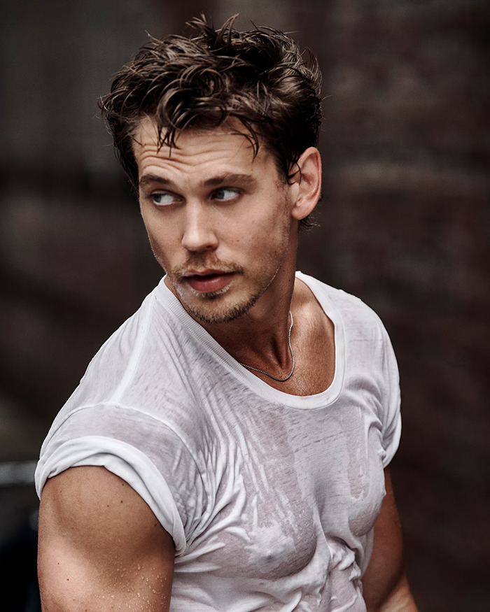 Austin Butler in a shirtless photoshoot showcasing his peak male body with wet hair and a white wet t-shirt against a dark background.