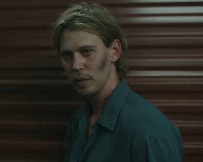 Young man with bruises on his face, wearing a dark shirt, posing against a brown striped background in dim light, peak male body.
