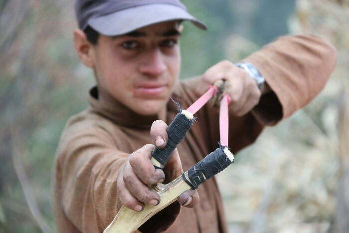 Boy outdoors wearing a cap and brown shirt pulling back a slingshot focused on learning new facts and curious activities