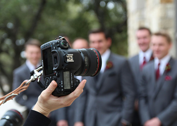 Hand holding a professional wedding camera capturing group of groomsmen in gray suits during outdoor wedding photoshoot.