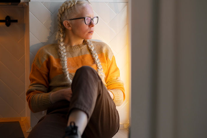 Woman with glasses and braided hair sitting by a window, reflecting on family conflict about disabled daughter and pregnancy.