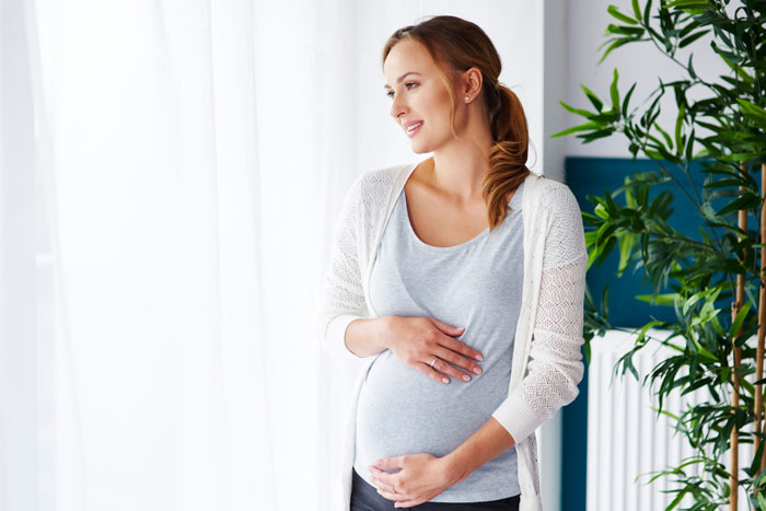 Pregnant woman in casual clothes gently holding belly indoors, representing parents obsessed with having kids.