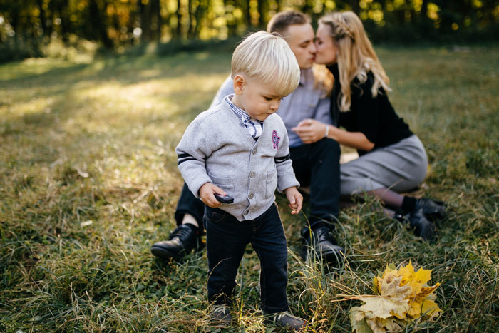 Young boy standing in a grassy field while parents sit behind, highlighting parents obsessed with having kids and older son babysitting.