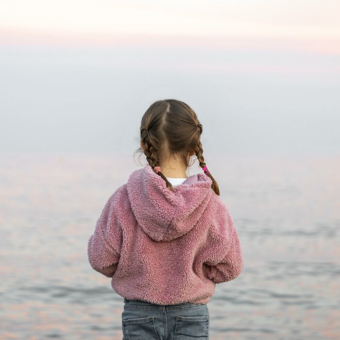 Child with braided hair and pink fuzzy hoodie standing by the water, capturing a moment of quiet reflection on Reddit fears.