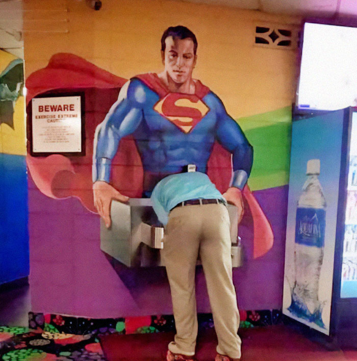 Man drinking from a water fountain integrated into a mural of Superman, illustrating a creative design fail.