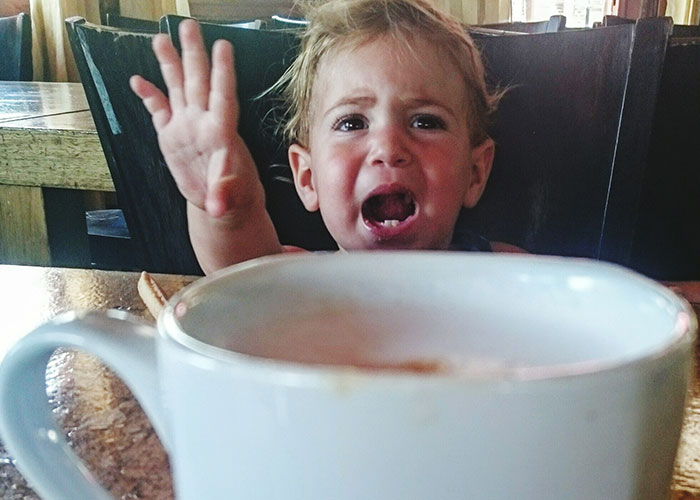 Toddler reaching out with an unhinged expression while sitting at a table behind a large coffee cup in a chaotic setting.