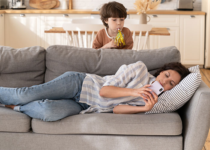 Child blowing party blowers behind a mom resting on a couch, showcasing unhinged mom hacks balancing genius and chaos.