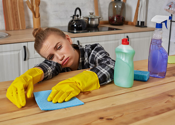 Tired mom wearing yellow gloves cleaning kitchen counter with cloth and cleaning supplies showing exhausted expression.