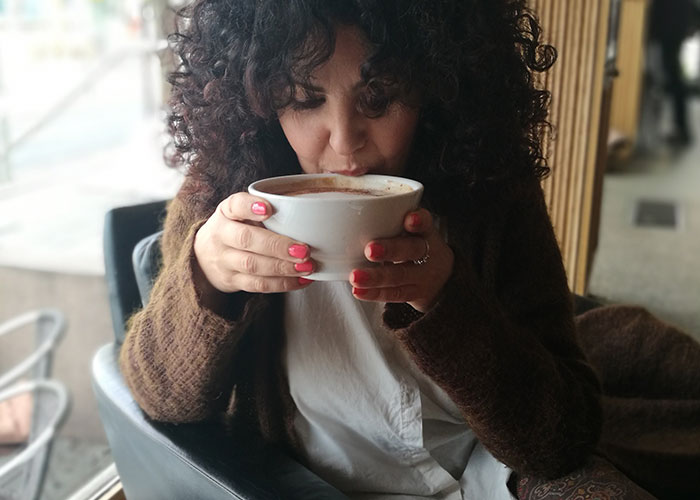 Woman with curly hair sipping a large cup of coffee at a café, highlighting unhinged mom hacks that toe the line between genius and chaos.