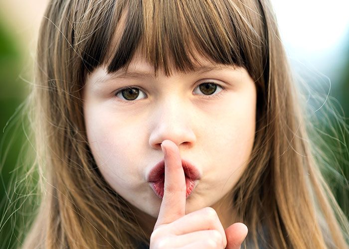 Young girl with long hair holding finger to lips in a quiet gesture, highlighting unhinged mom hacks between genius and chaos.