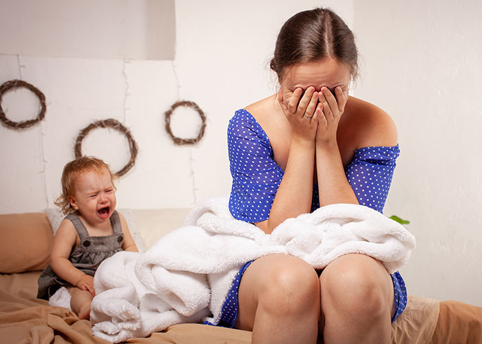 Frustrated mom covering her face while toddler cries in the background, reflecting unhinged mom hacks chaos and genius.