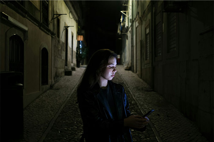 Young woman using smartphone alone on a dark alley at night, illustrating street smart tips and tricks for safety.