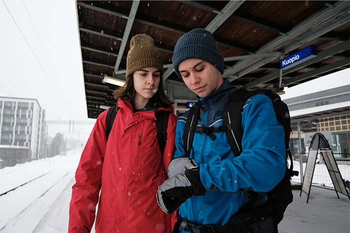 Two street smart travelers in winter gear at a snowy train station, checking time and planning their next move.