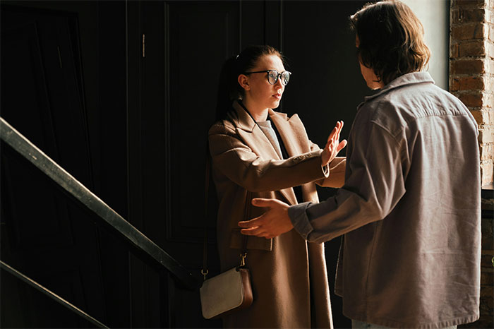 Woman using street smart communication skills, wearing glasses and coat, gesturing clearly while talking to a man indoors.