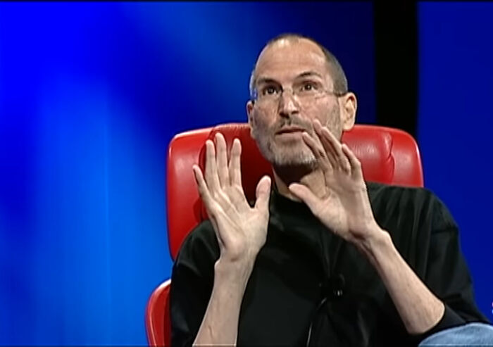 Man in black shirt seated on red chair, speaking with expressive hand gestures about biggest world unexpected discoveries.