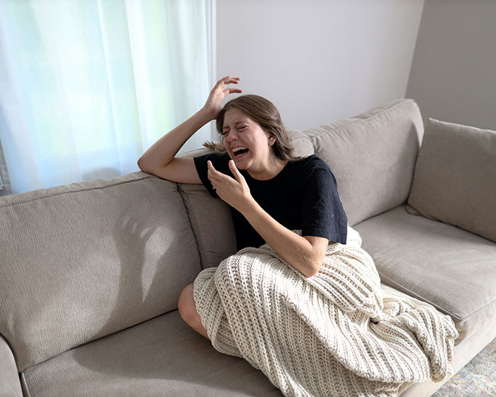 Young woman crying on couch wrapped in blanket expressing distress, relating to heavily pregnant woman stuck in bathtub scenario.