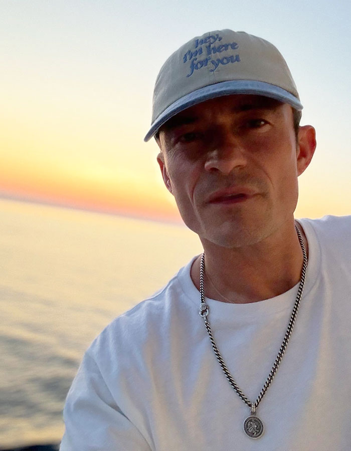 Orlando Bloom wearing a cap and necklace, posing near water during sunset, trolling Katy Perry and Justin Trudeau rumors.