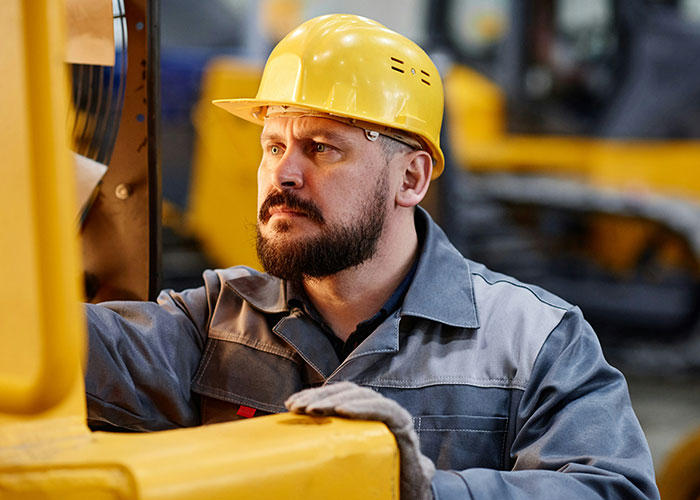 Blue collar worker wearing a yellow hard hat inspecting heavy machinery at a job site focusing on safety risks.