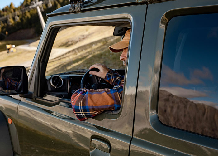 Blue collar worker wearing a cap and plaid shirt, driving a green vehicle near a hillside worksite on a sunny day.