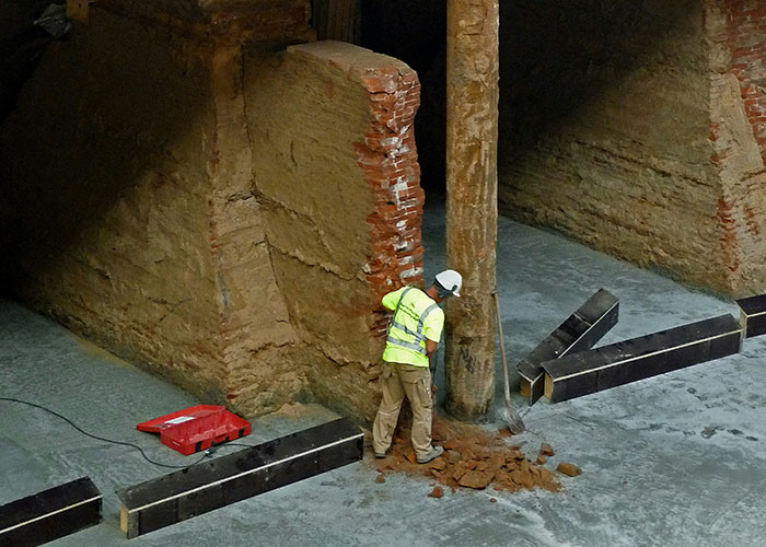 Construction worker in safety gear clearing debris from a damaged pillar in a job site with exposed brick walls.