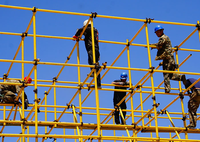 Blue collar workers wearing helmets working on yellow scaffolding at a job site under a clear blue sky.