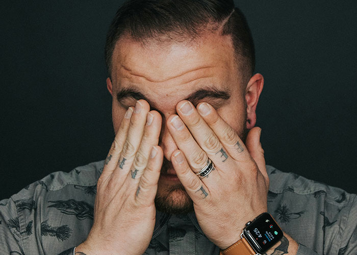 Blue collar worker with tattoos and smartwatch covering his face, reflecting on a scary job site experience.