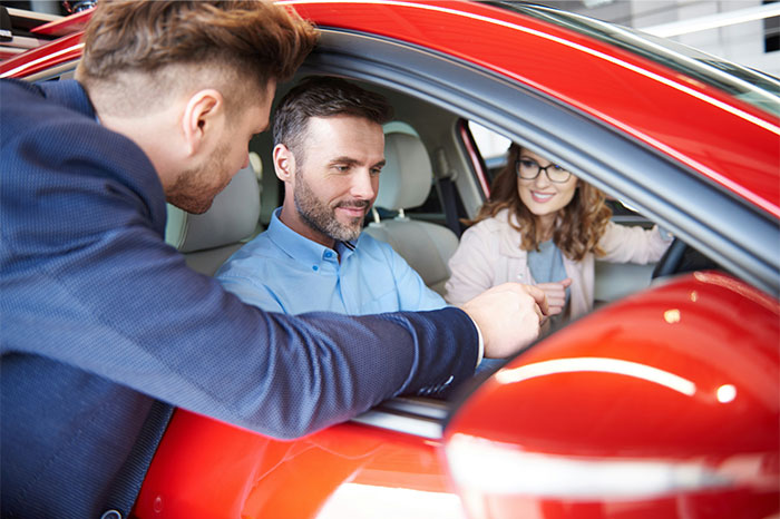 Car sales professional showing key to a couple inside a red car, illustrating profitable industries today.