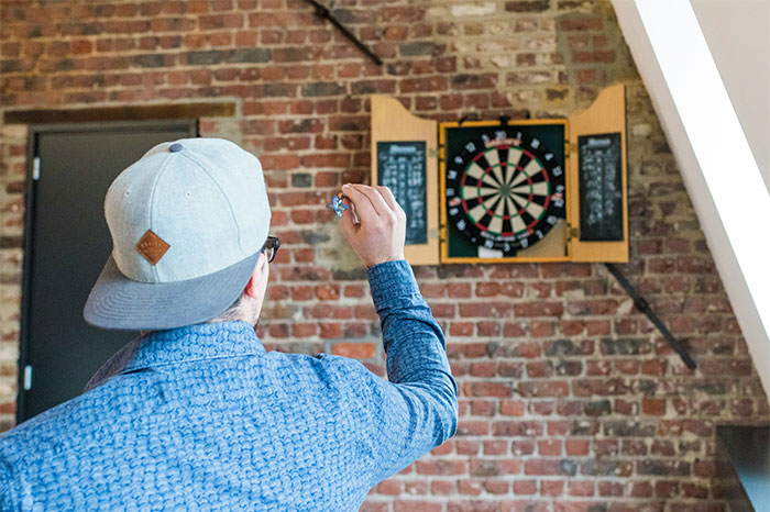 Man wearing a cap aiming darts at a board in a brick-walled room representing profitable industries concept.