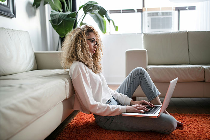 Young woman with curly hair and glasses sitting on floor working on laptop, representing profitable industries these days.