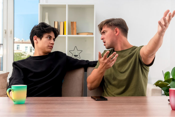 Two young men arguing at a table inside a home, showing tension between a boyfriend and a roomie over chores.