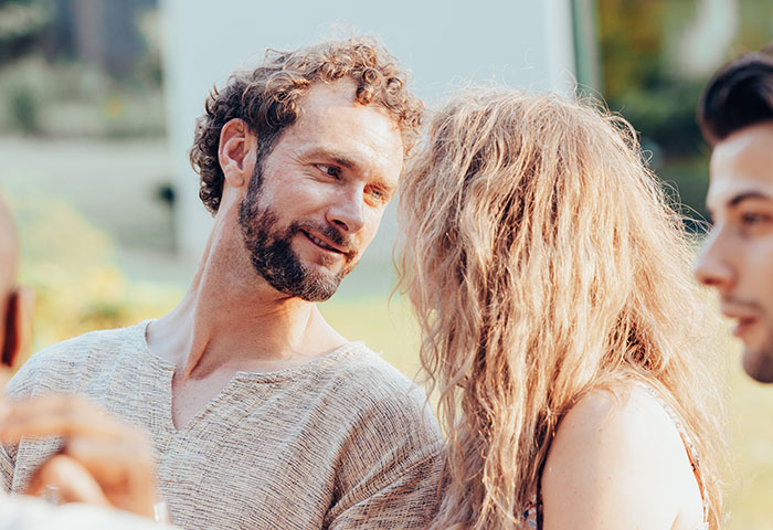Man pretending to be single at a party talking to a woman while another man looks on nearby outdoors.